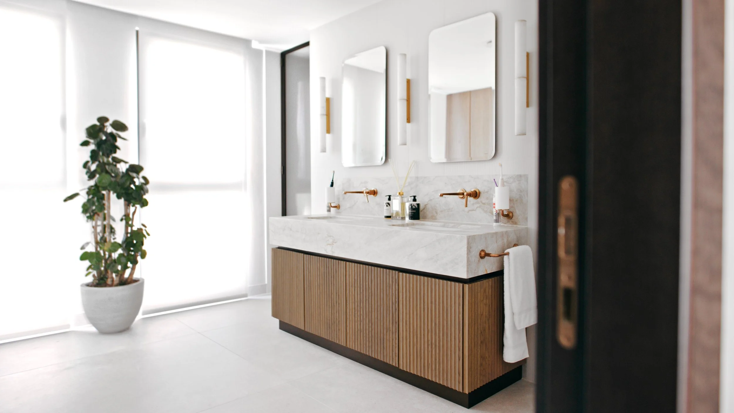 Modern bathroom with a double marble sink vanity, gold fixtures, three mirrors, a potted plant, and large windows with blinds.