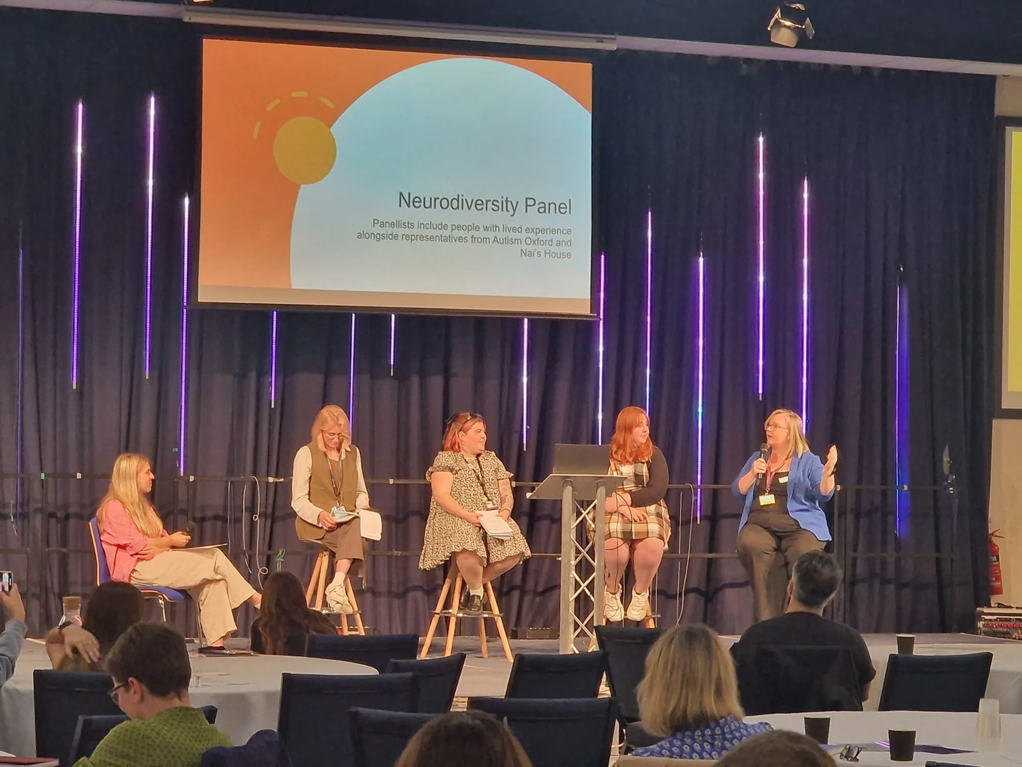 A panel discussion at a conference called Neurodiversity Panel, with five women on stage, one speaking into a microphone. Behind them is a large presentation screen with the title of the panel.