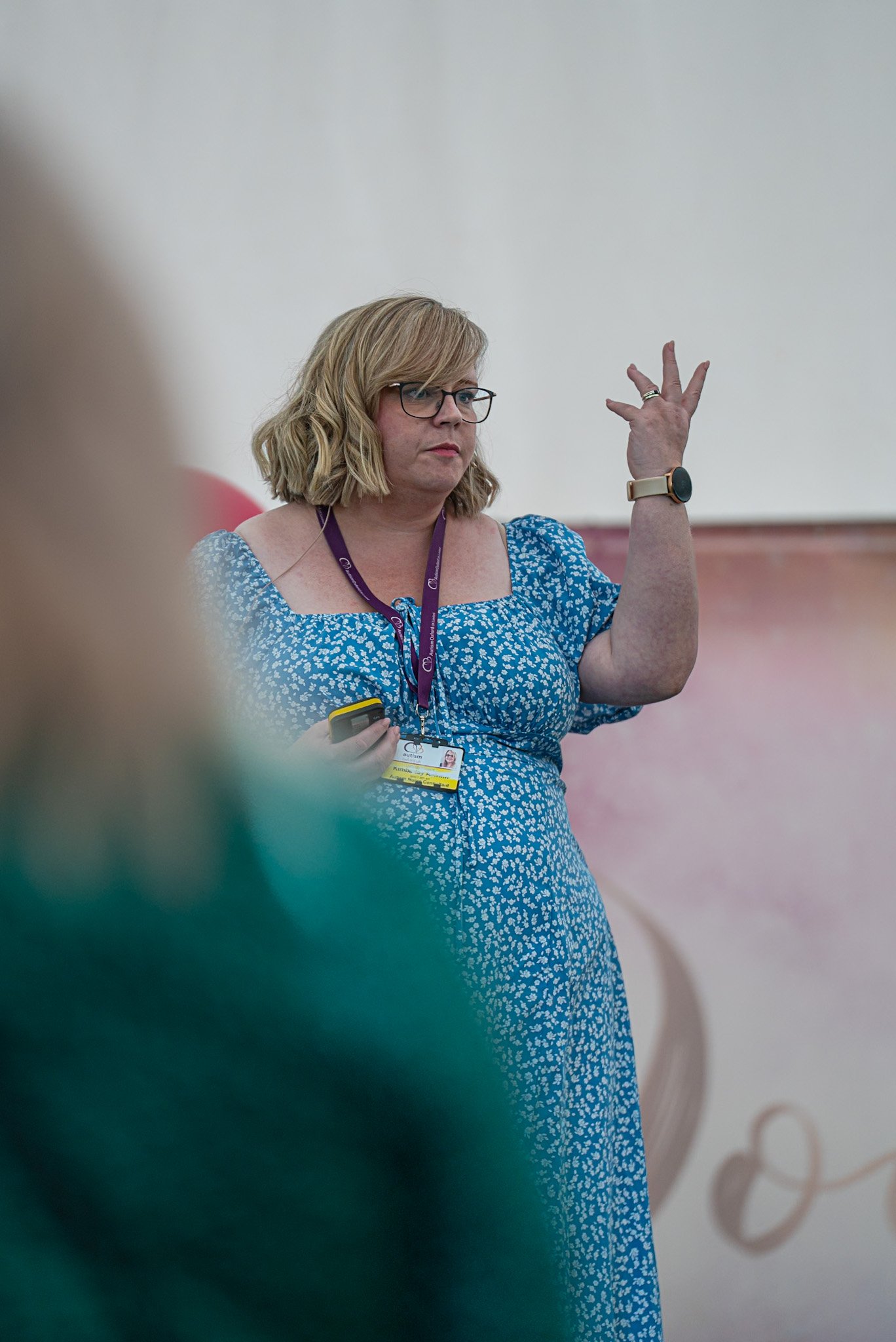 A woman with blonde hair, glasses, and a blue dress with a white floral pattern stands in front of a beige wall, holding a yellow remote and wearing a purple lanyard with an ID badge. She has a serious expression and is gesturing with her left hand.