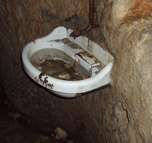 An old, rusted toilet bowl mounted against a rough, uneven stone wall, with visible rust and grime.