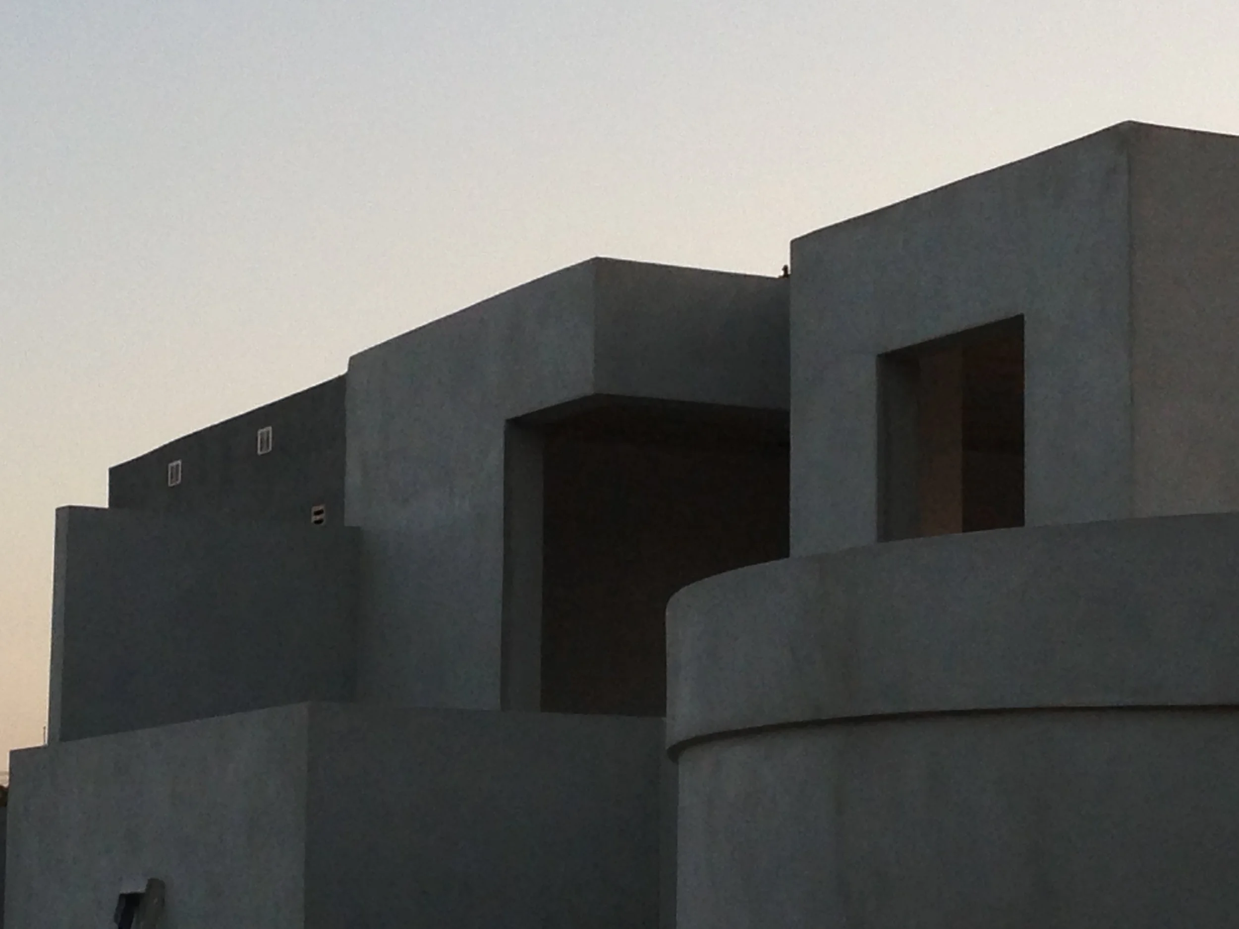 Close-up view of a modern building with geometric design, gray exterior walls, and open window spaces, under a pale sky.