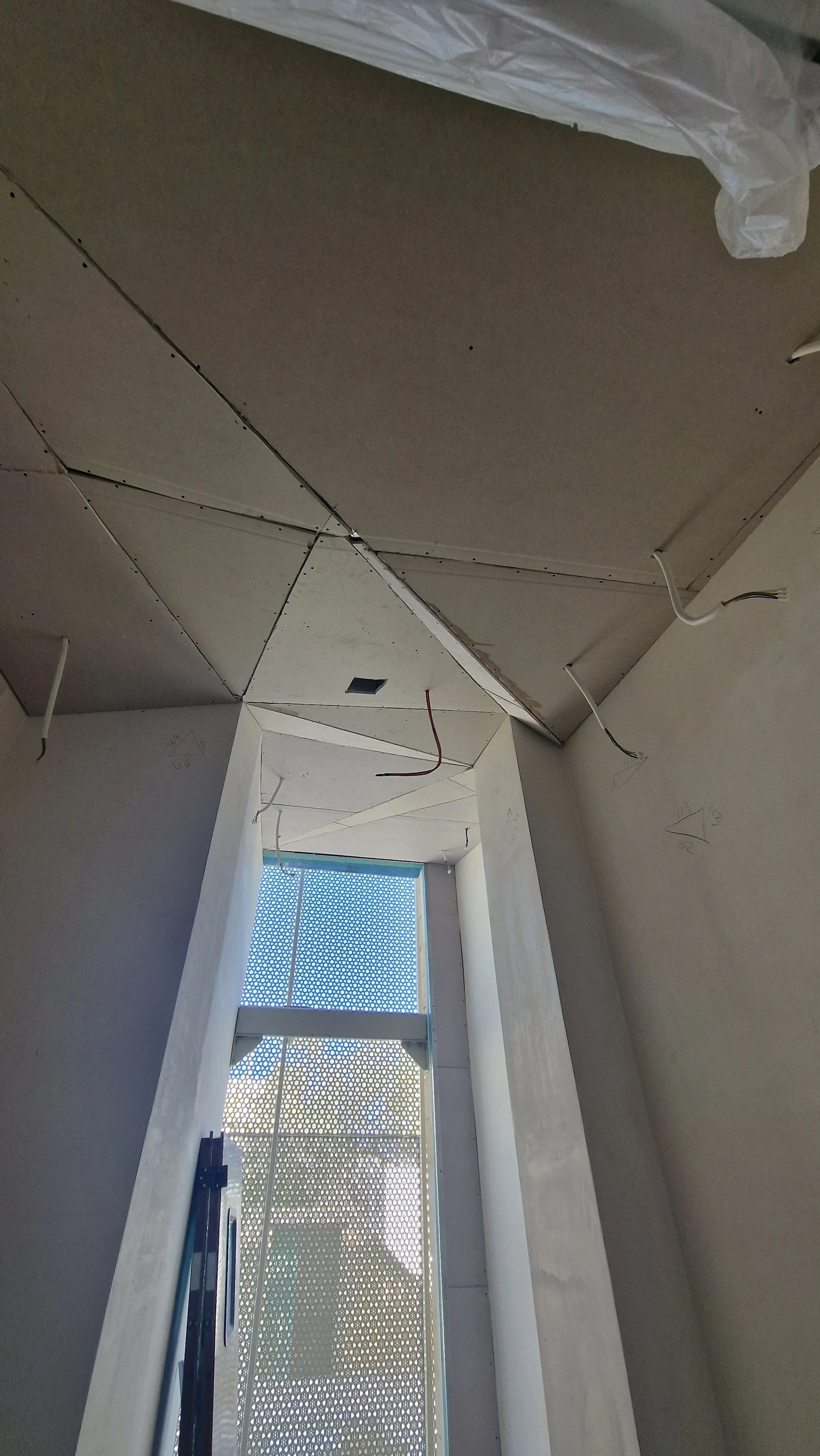 Interior room under construction with drywall ceiling, exposed wires, and a window with a perforated metal screen.