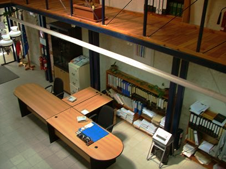 An office space with a large wooden desk, black chairs, shelves filled with binders and papers, a small filing cabinet, and background equipment.