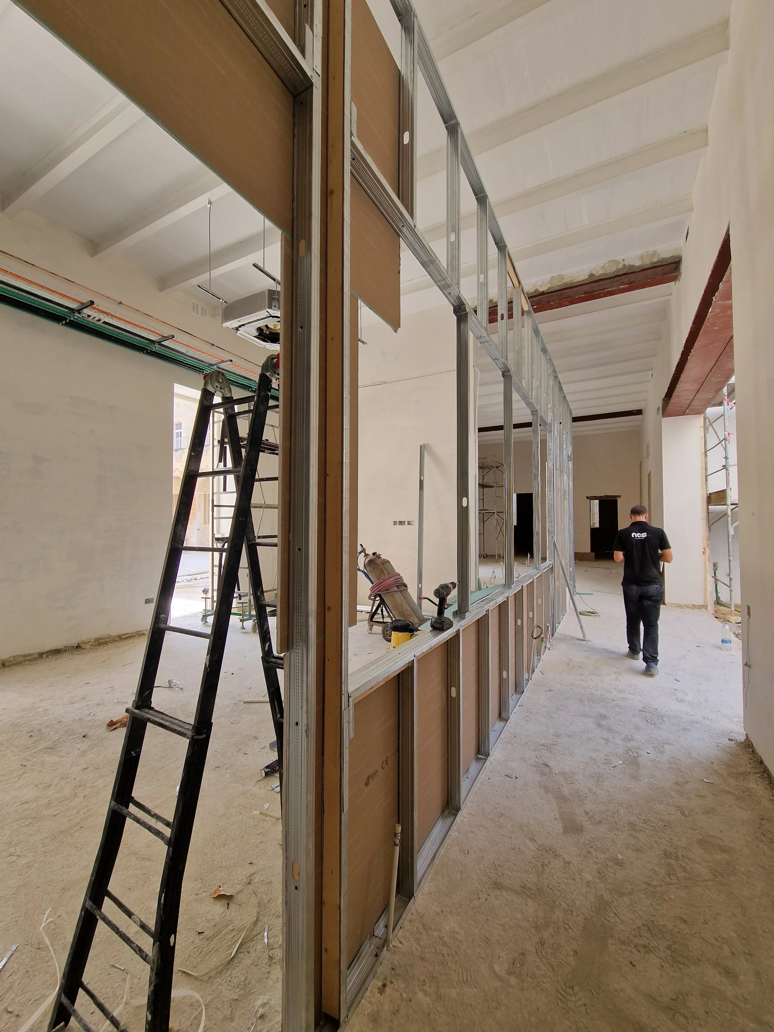 Interior construction site with metal framing and drywall panels, ladder, electrical wiring, and workers installing wall partitions.