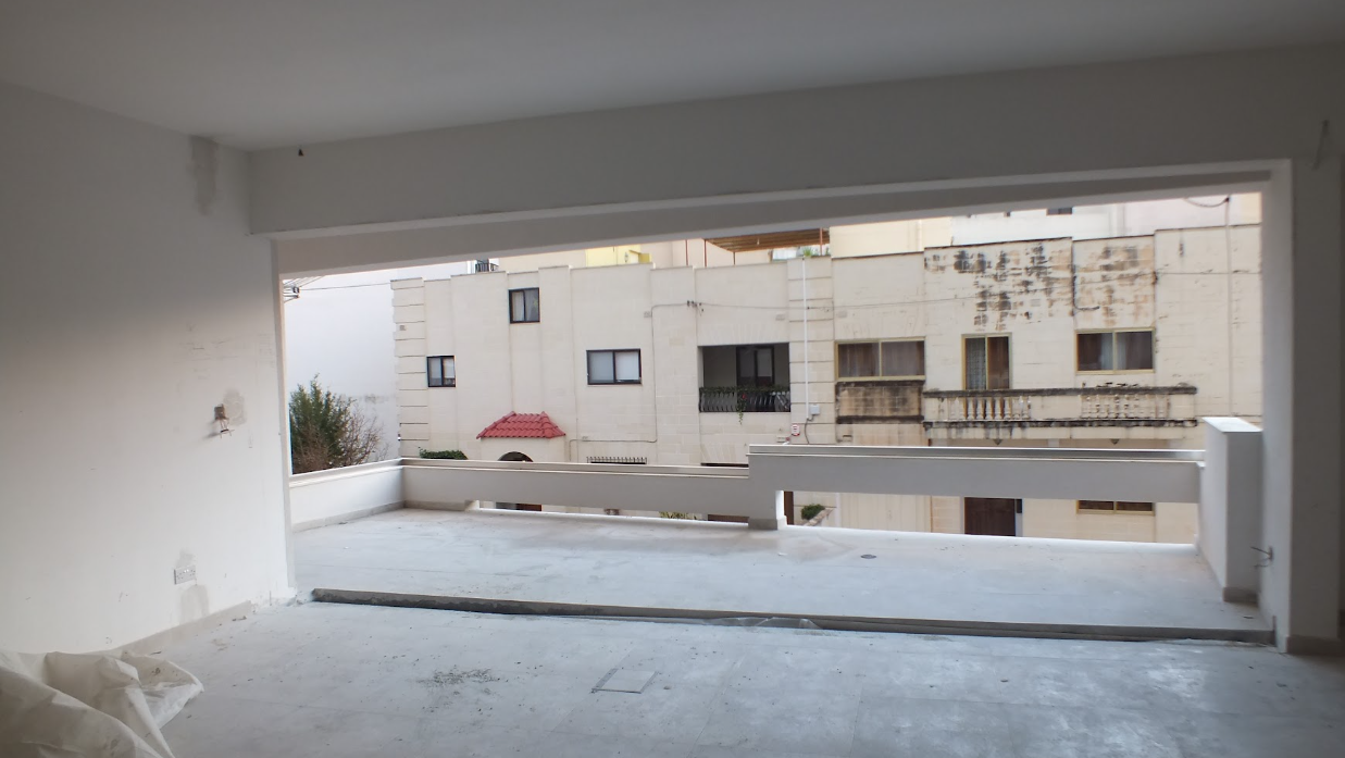 Unfinished apartment balcony with concrete floor and partial wall railing, overlooking neighboring buildings with white facades and small windows.