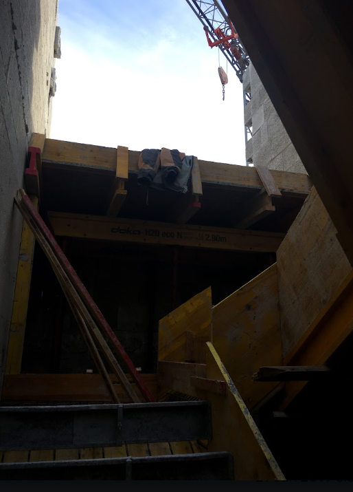 Construction site with wooden framing, ladders, and tools, with a crane and sky visible in the background.