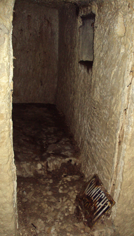A narrow stone passageway with rough, uneven walls and floor, and a metal barricade on the ground with the word 'CAUTION'.