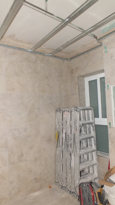 A room under construction with a ladder, drywall or plaster walls, a window, and an unfinished ceiling with metal framing.