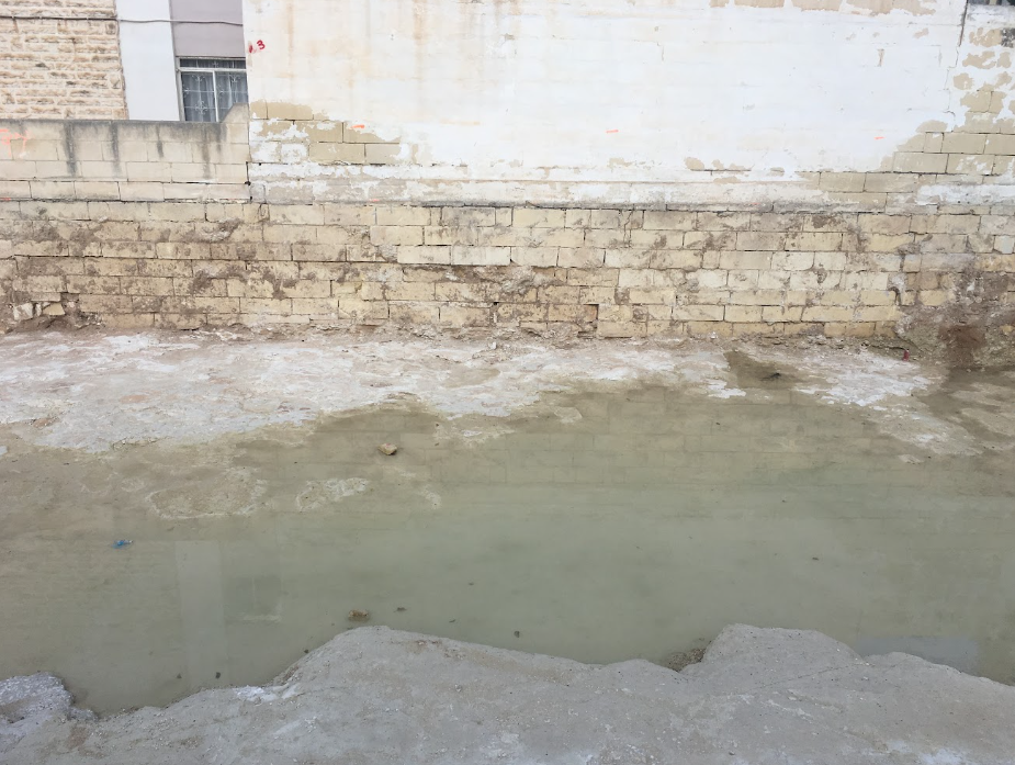 A waterlogged construction site with muddy water, brick wall, and partially built or damaged concrete ground.