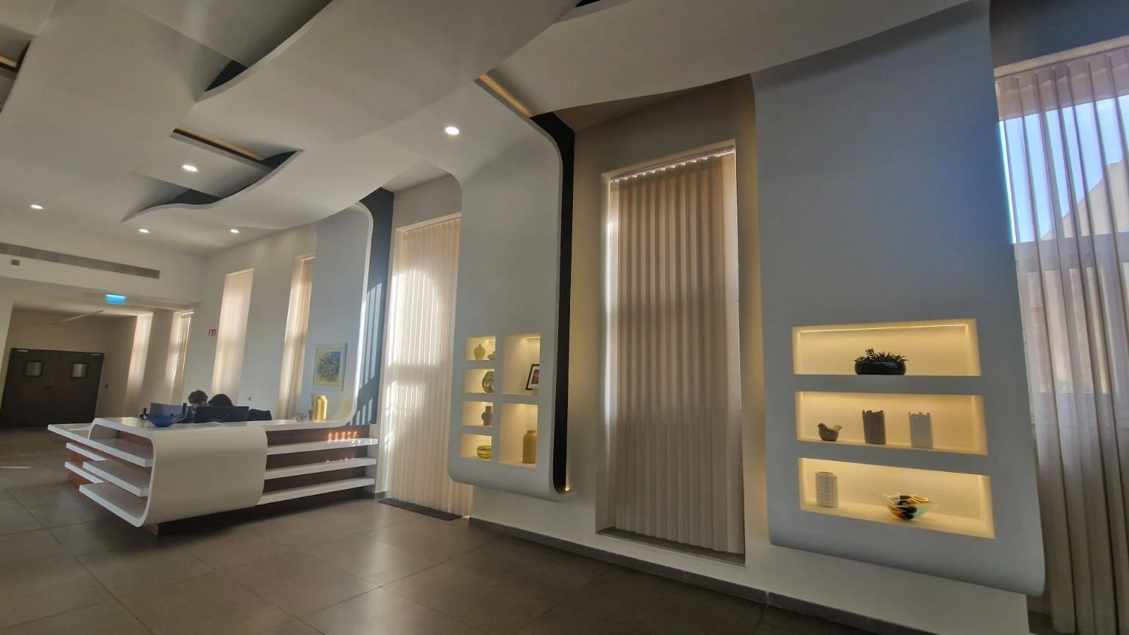 Modern interior lobby with white curved reception desk, tall windows with vertical blinds, and built-in illuminated shelves displaying decorative items.