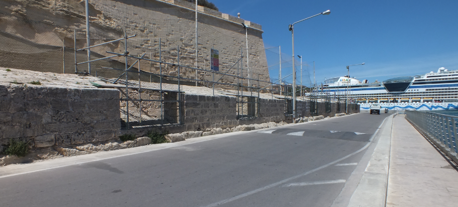 Empty road alongside a construction site with scaffolding, with large cruise ships docked in the background under a clear blue sky.