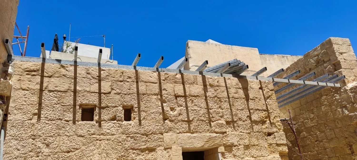 Construction site showing a stone wall with metal supports and scaffolding against a bright blue sky.