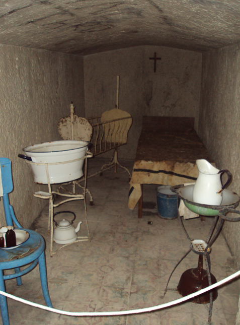 A small, rustic, underground room with a bed, an old brass bed, and an altar with a cross on the wall.