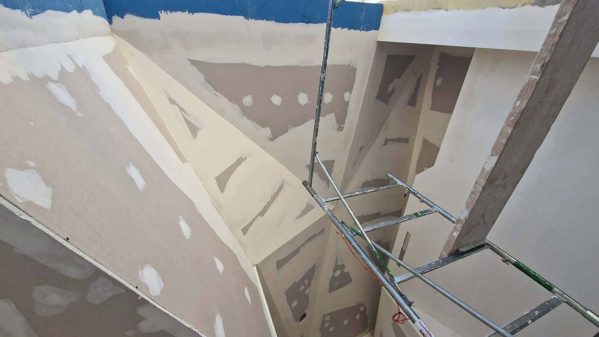 Interior view of a building under construction with drywall installation, scaffolding, and unfinished walls with patches of joint compound.