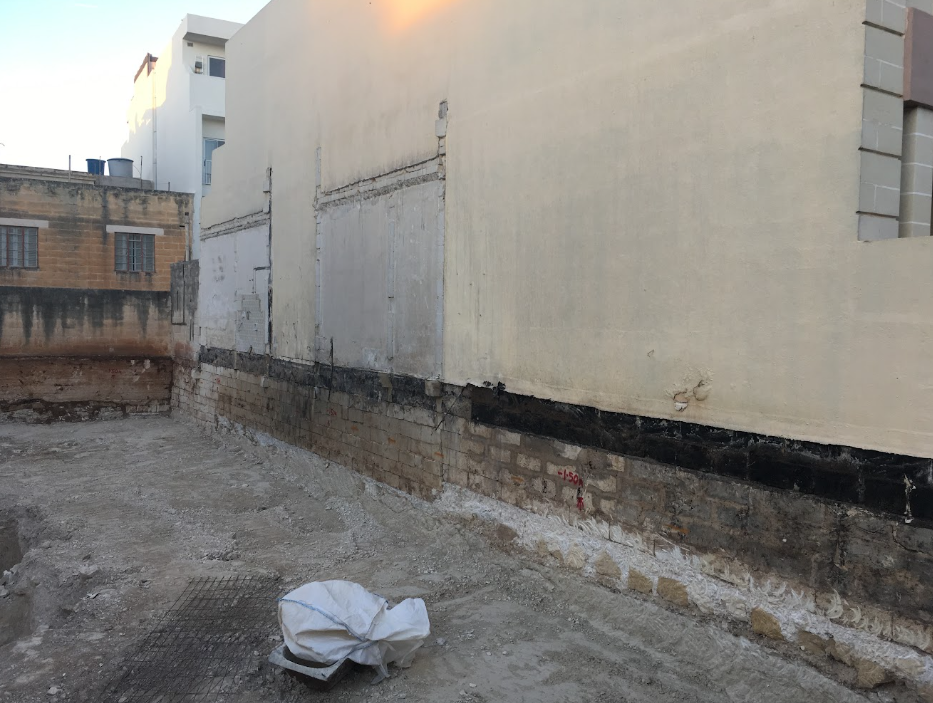 Construction site with exposed brick and concrete wall, and a bag of construction materials on the ground.