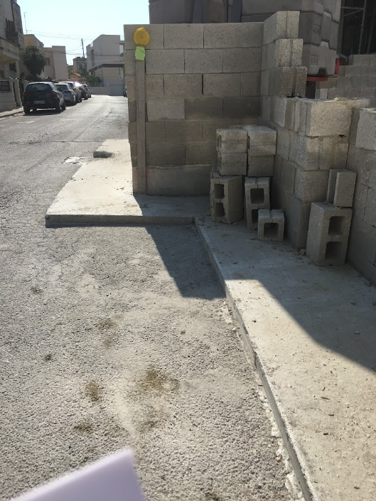 Cinder block wall under construction on a sidewalk with scattered cinder blocks, a yellow tape measure, and shadow cast on the ground.