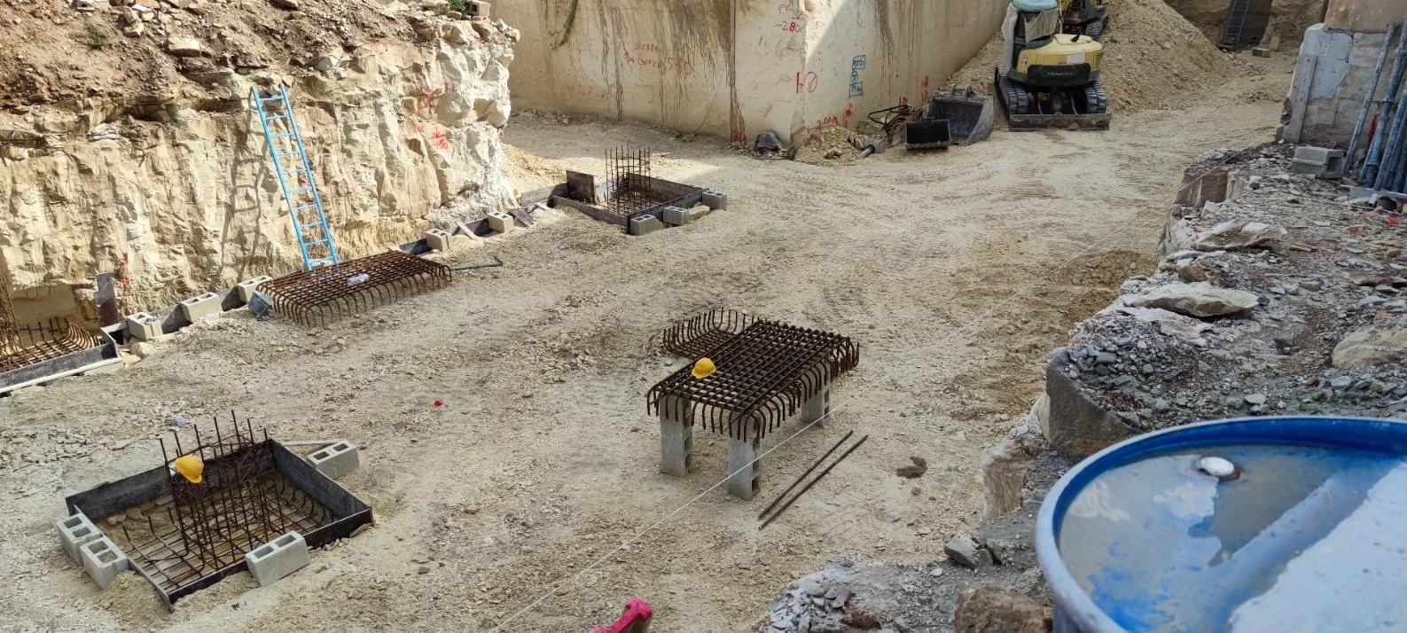 Construction site with reinforced steel rebars, concrete block foundations, a ladder leaning against a rocky wall, and construction equipment in the background.