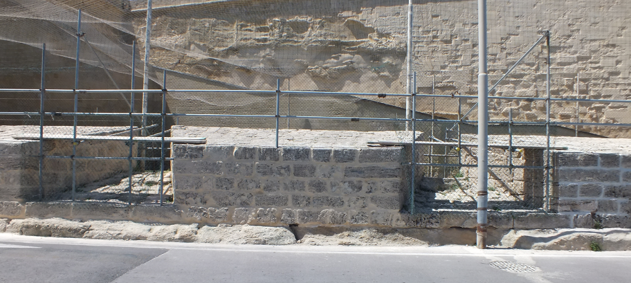 Stone retaining wall with metal railing and scaffolding along a sidewalk in front of a eroded hillside or cliff.