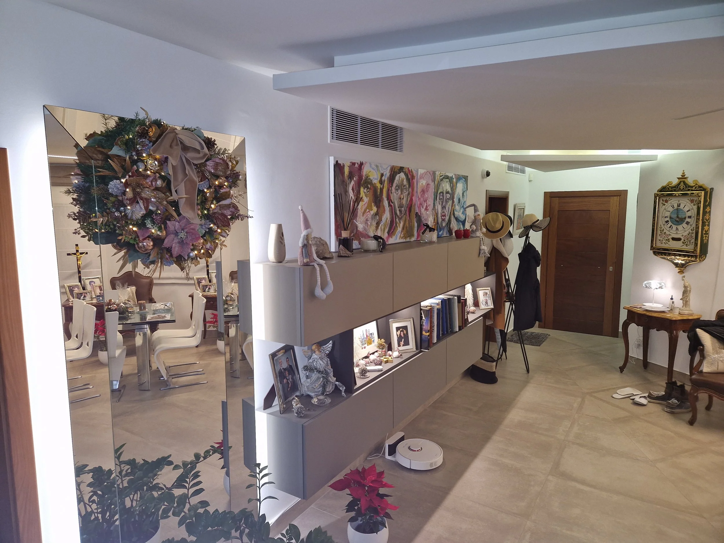An indoor living space with a decorated Christmas tree, a white cabinet with framed photos, artwork of faces on the wall, hats hanging, a clock, and household items.