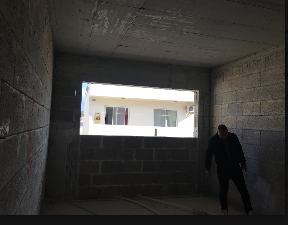 Interior view of an unfinished building with concrete walls and ceiling, with a large rectangular opening revealing an exterior balcony with a white building in the background; a person is standing inside, silhouetted against the bright outside.