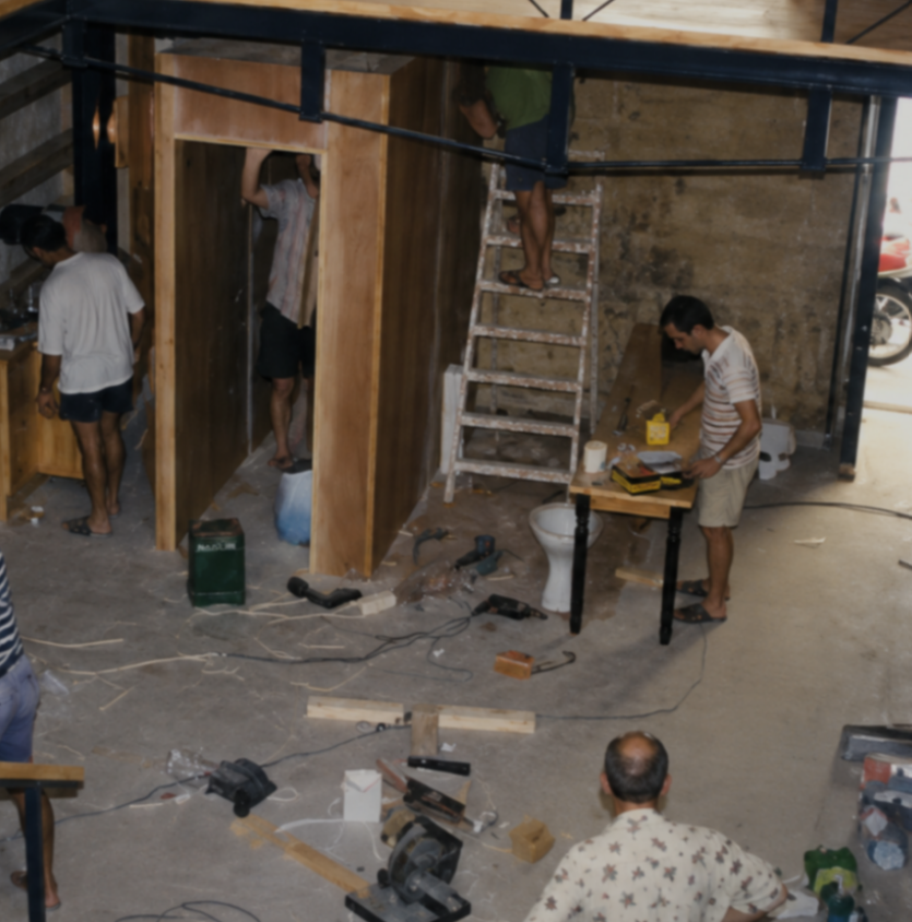 People working on building or renovating a small wooden structure indoors with tools and construction materials scattered on the floor.