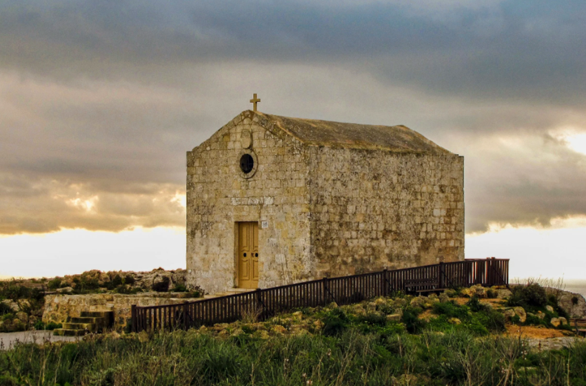 Stone church with a cross on top, adjacent to a black fence, under a cloudy sky, surrounded by grass and rocks.