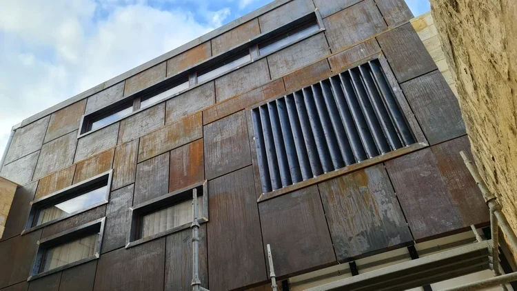 Close-up of a modern building exterior with rusted metal panels, rectangular windows, and a large vent with vertical black slats.
