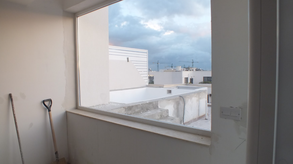 View from inside a room looking out a large rectangular window at an urban cityscape with white buildings and construction cranes under a cloudy sky.