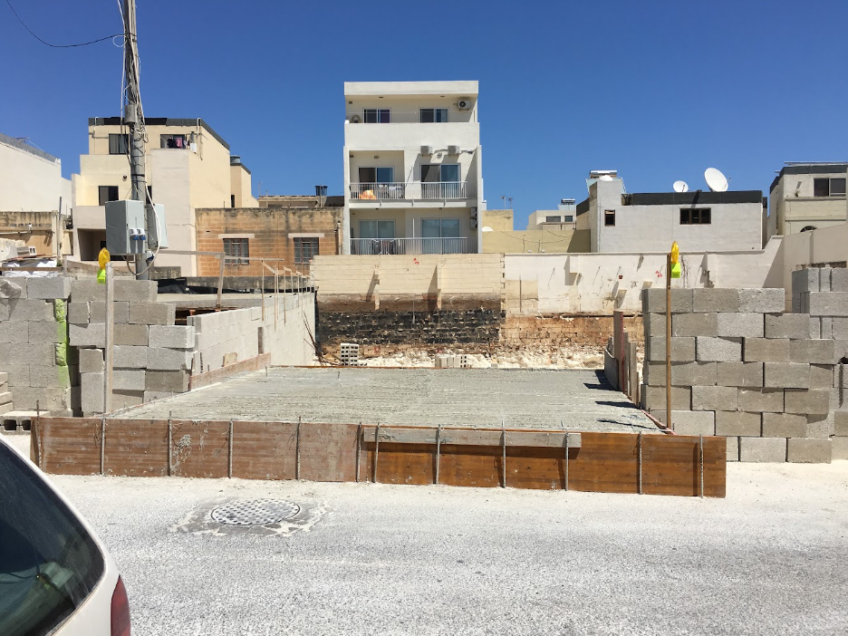 Construction site with concrete foundation and cinder block walls, surrounded by urban buildings and a clear blue sky.