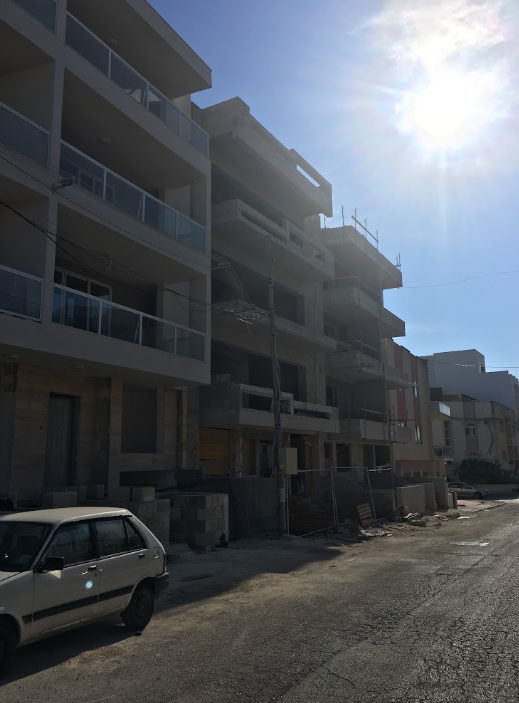 Under construction multi-story building with balconies, parked car on an unpaved street, bright sun in the sky.