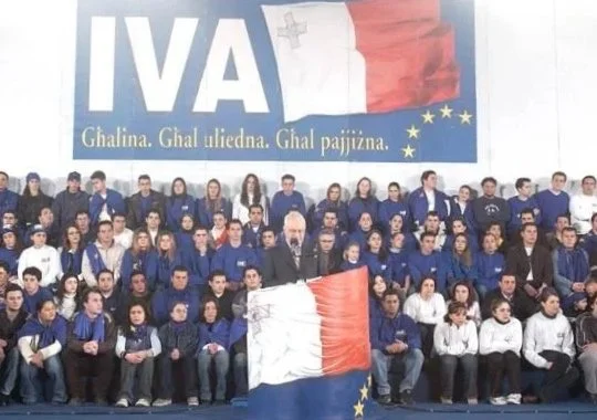 Group of young people seated on stage, wearing blue and white clothing, with a large banner behind them displaying the Italian flag, the word 'IVA,' and European Union stars.