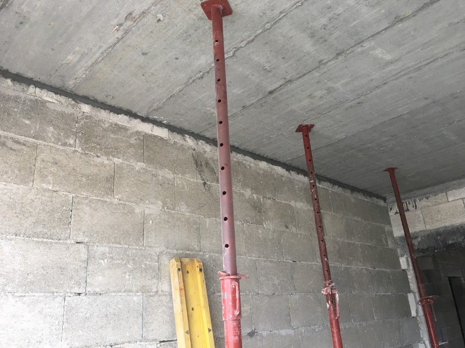 Construction site with three red adjustable steel support poles holding up the concrete ceiling, and a yellow wooden ladder leaning against a gray cinder block wall.