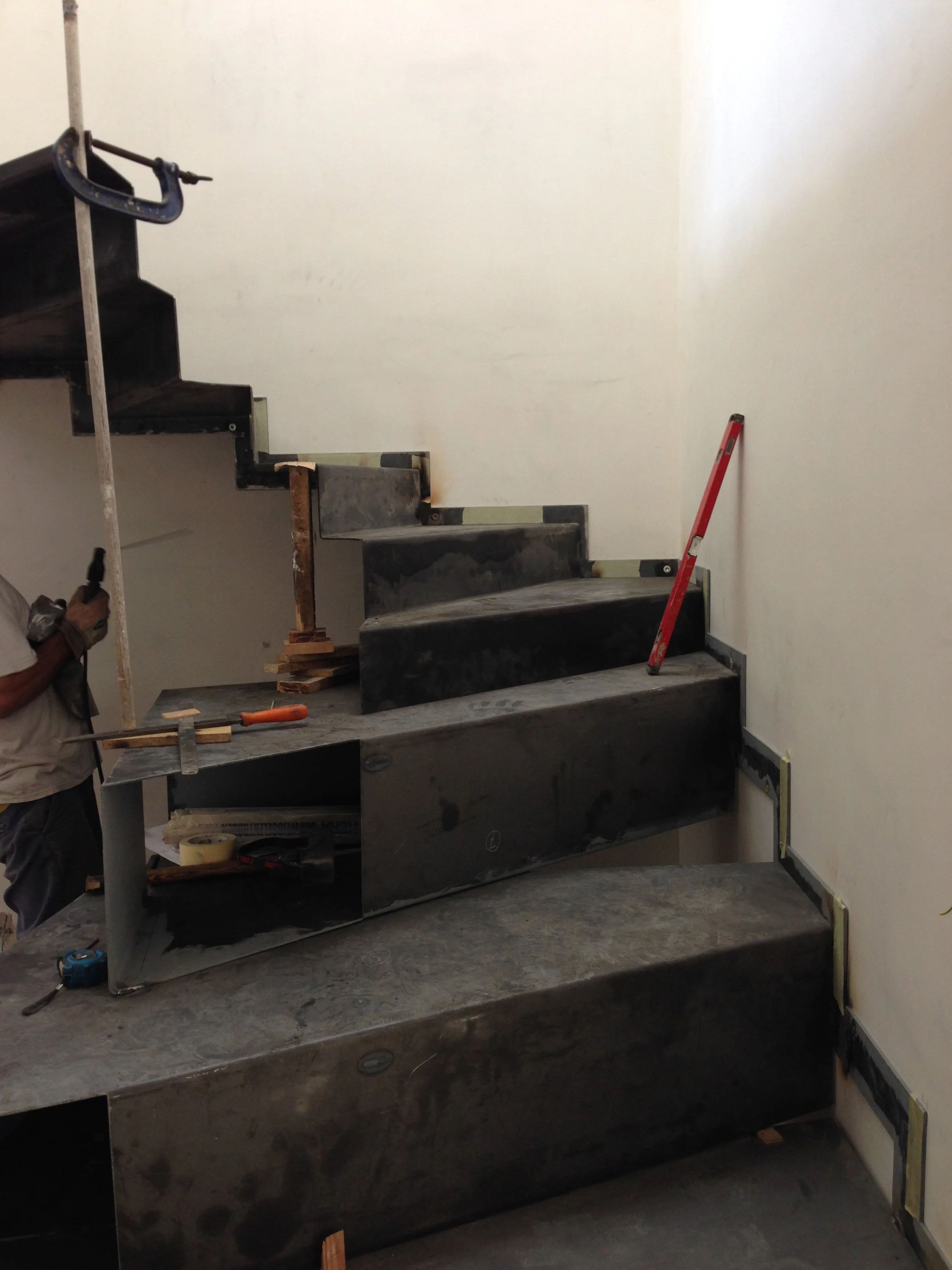 Under construction staircase with black steps, a red level tool leaning against the wall, and construction equipment and tools nearby.