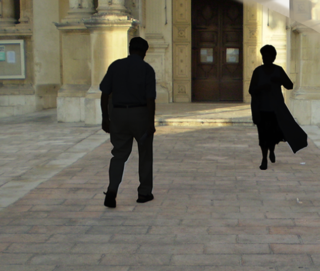 Two people walking away from the camera outside a historic building with stone architecture and large wooden doors.