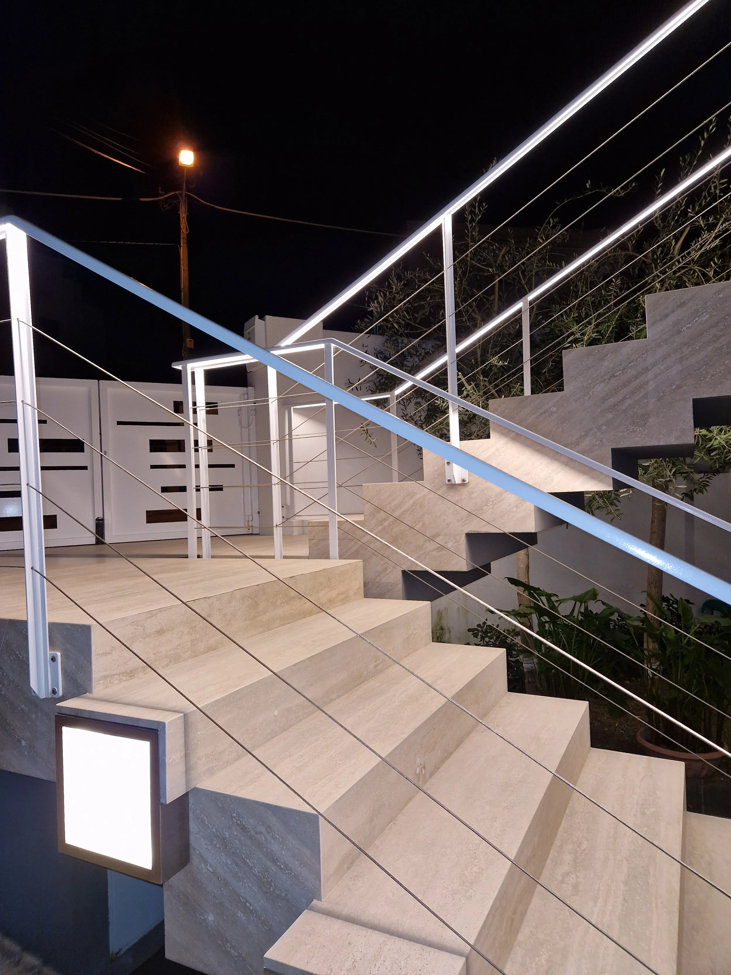 Night view of an outdoor staircase with beige stone steps and white metal handrails, illuminated by strips of LED lights, next to a modern building with a white fence, greenery, and a lamp post in the background.