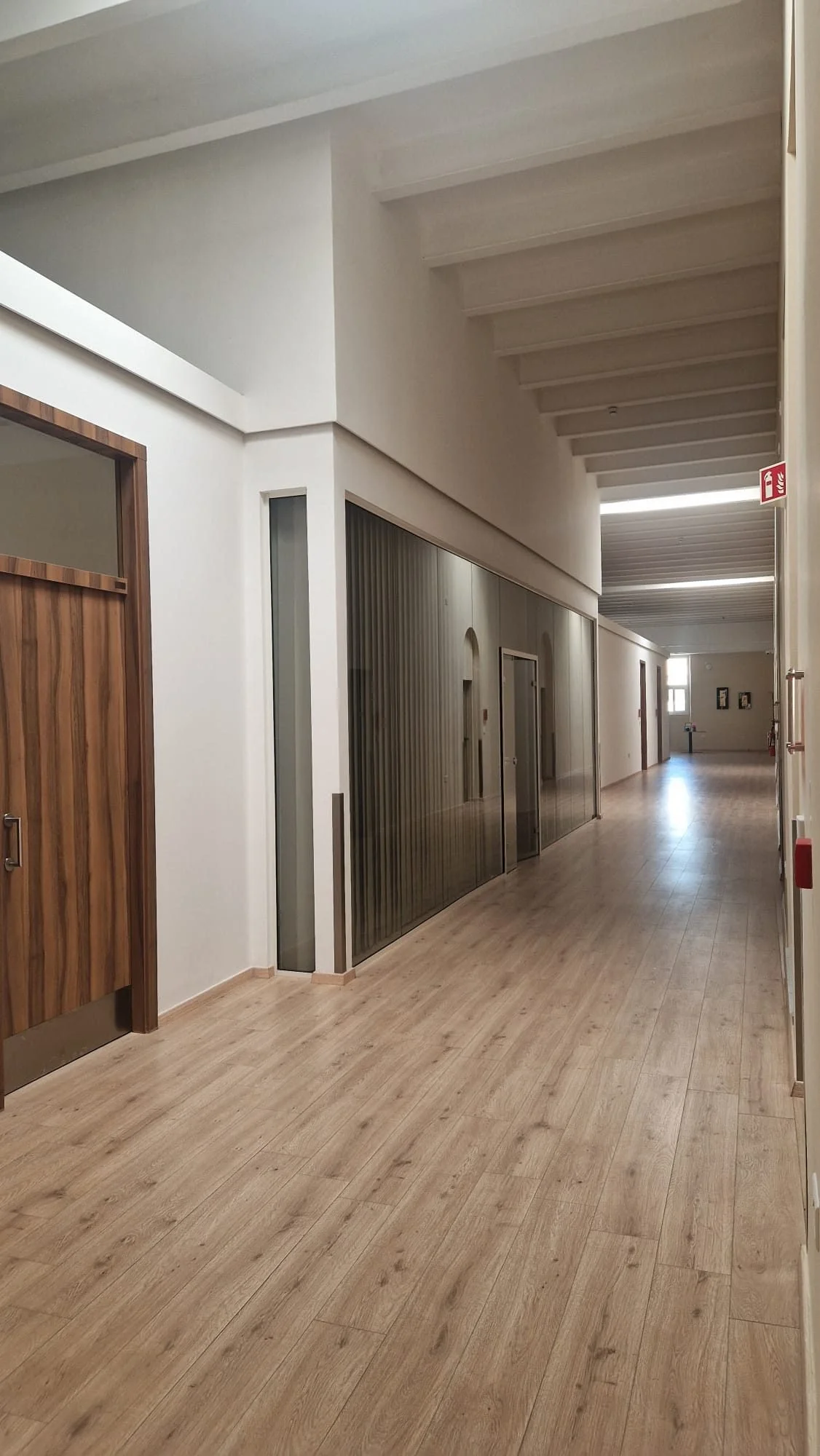Empty corridor with wooden flooring, white walls, ceiling with exposed beams, wooden doors on the left, and a fire extinguisher sign on the right.