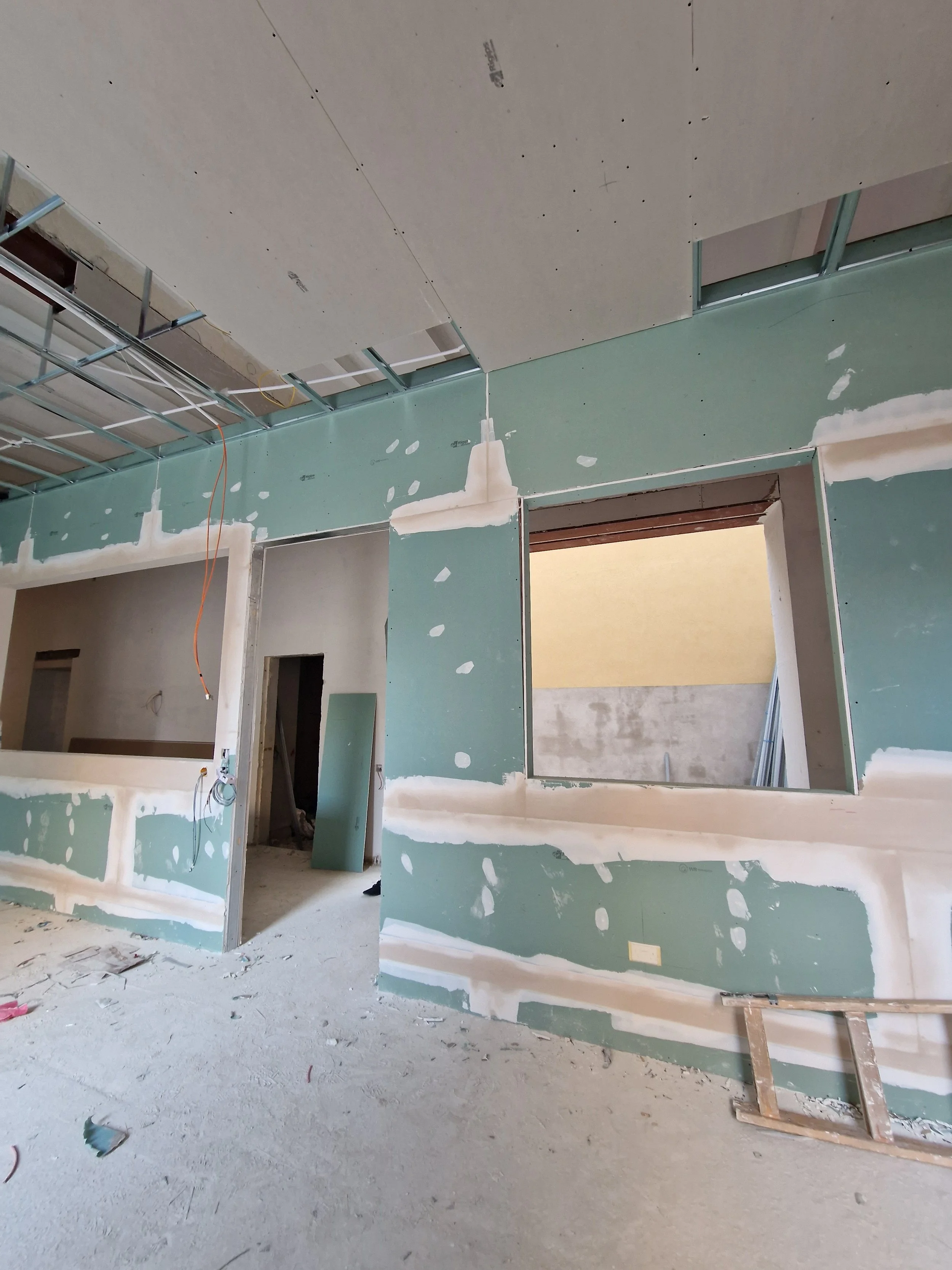 An indoor construction site with drywall installation in progress, exposed metal framing, and unfinished walls and ceiling.