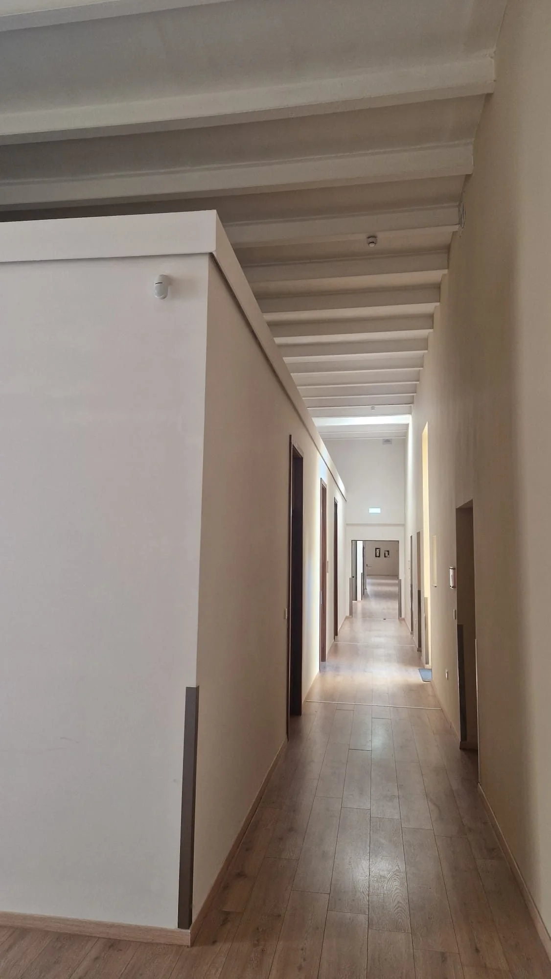 A long hallway with beige walls, wooden flooring, and a high ceiling with exposed beams. Several doorways are visible along the hallway, leading to different rooms, with some framed pictures on the walls at the far end.