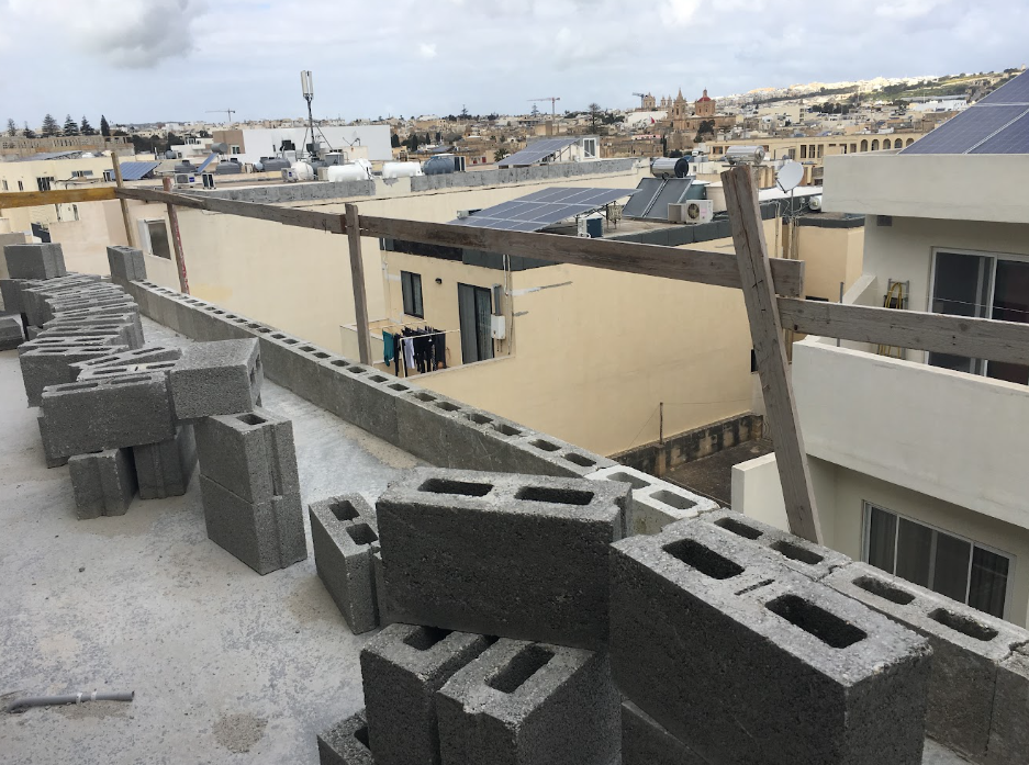 Construction site on a building rooftop with stacked cinder blocks, wooden support beams, and neighboring buildings with solar panels and clothing hanging outside.