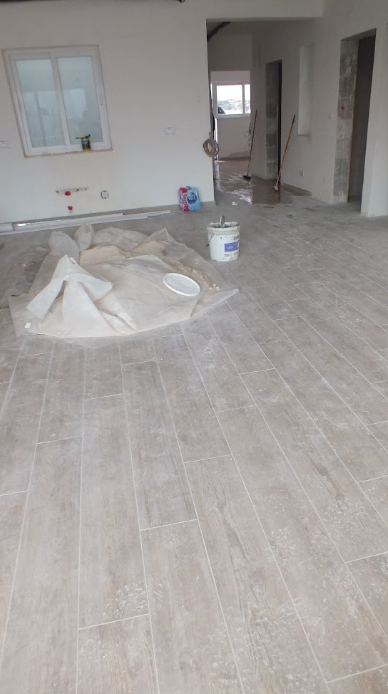 Interior of a house under construction or renovation, with beige tiled flooring, a white wall with a window, and construction materials including a tarp, a paint bucket, and cleaning supplies.