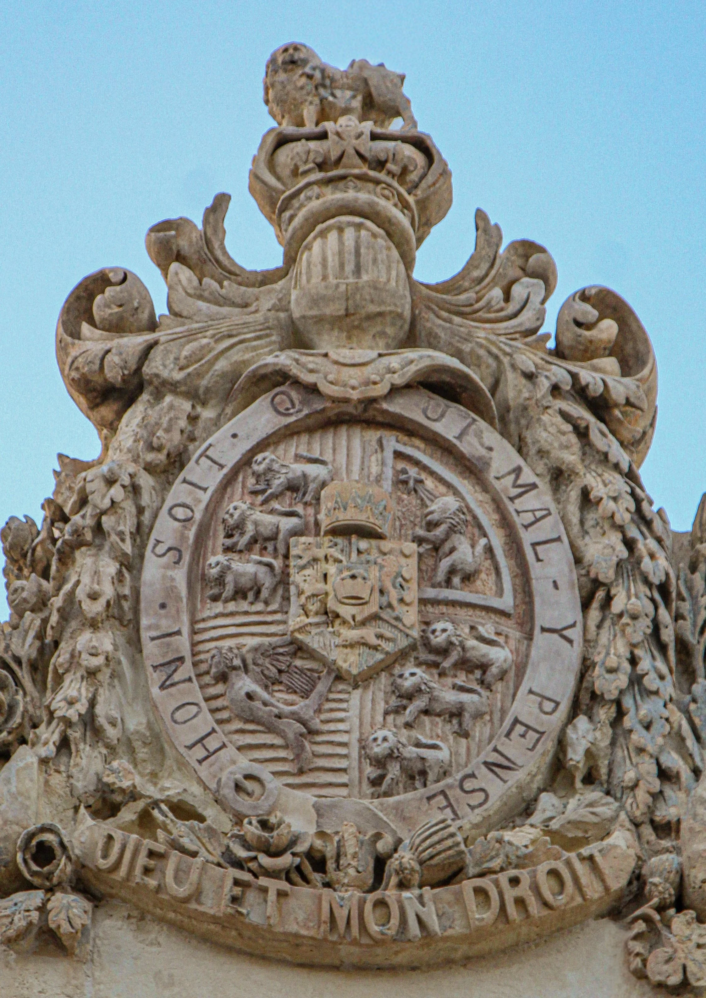 A historical building with a decorative stone crest featuring a lion on the left, a unicorn on the right, and a shield in the center. Below, a Latin inscription is carved into the stone, and a clock with black hands and Roman numerals is positioned beneath the inscription.