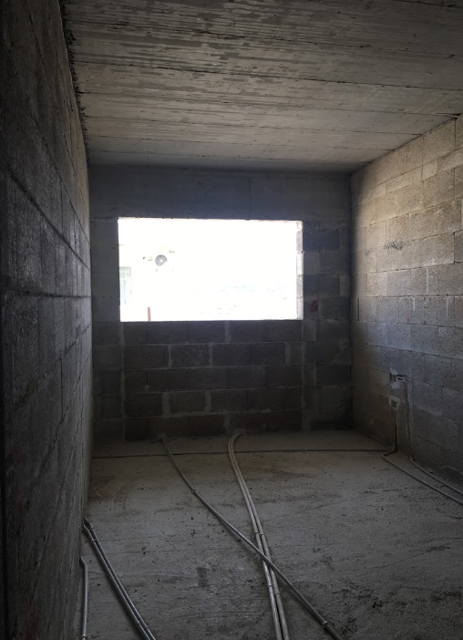 Interior of a building under construction with unfinished concrete walls and ceiling, a rectangular opening on the wall, and electrical wiring on the floor.