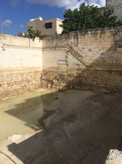 Empty outdoor swimming pool with concrete and stone walls, surrounded by buildings and greenery under a blue sky.