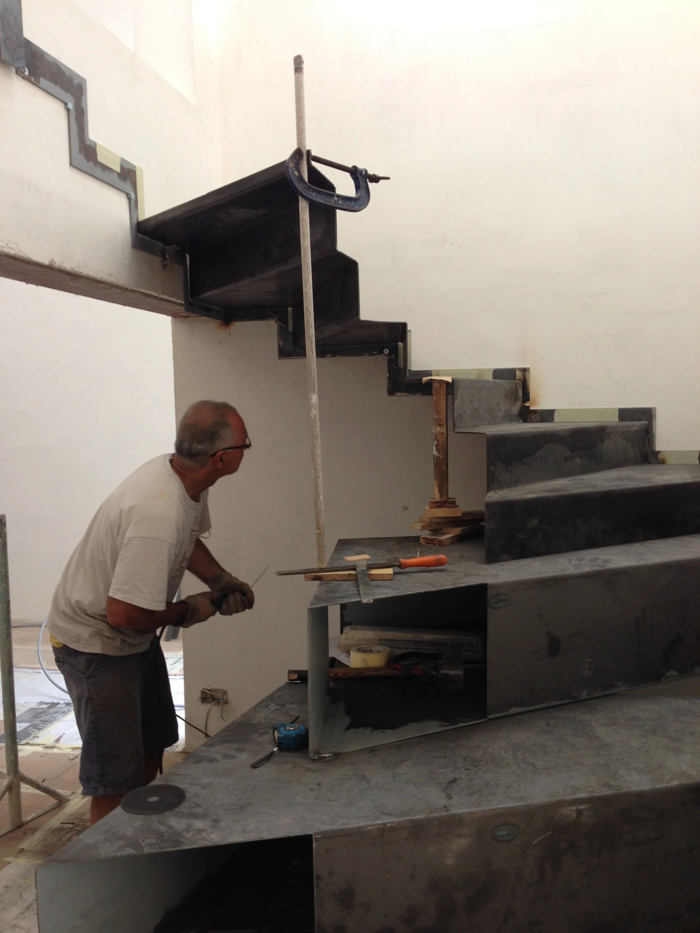 Construction worker standing near a large stone staircase, working on the stairs with tools. The staircase is partially built, with some sections unfinished and materials scattered around.