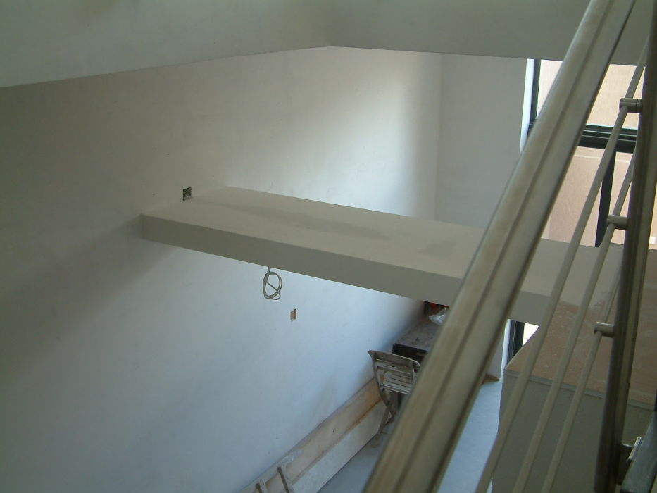 Interior view of a building under construction, showing a white painted wall, a suspended white beam, an electrical outlet, and part of a ladder or scaffolding.