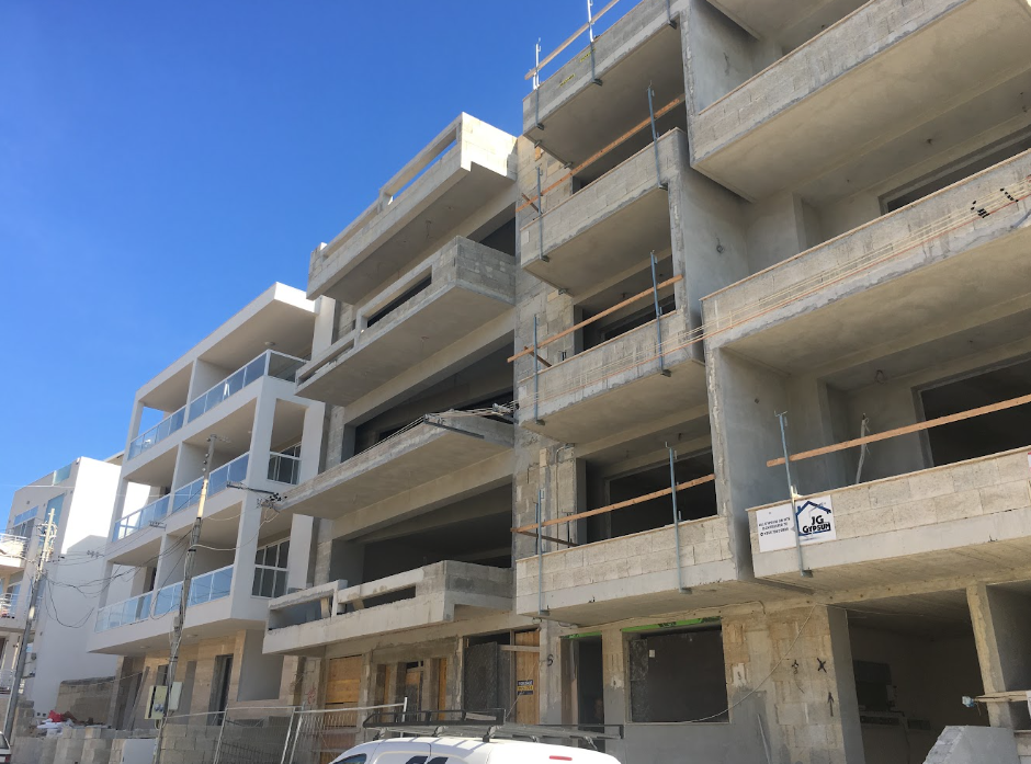 Under-construction multi-story residential building with exposed concrete and glass balconies, scaffolding, and a clear blue sky.