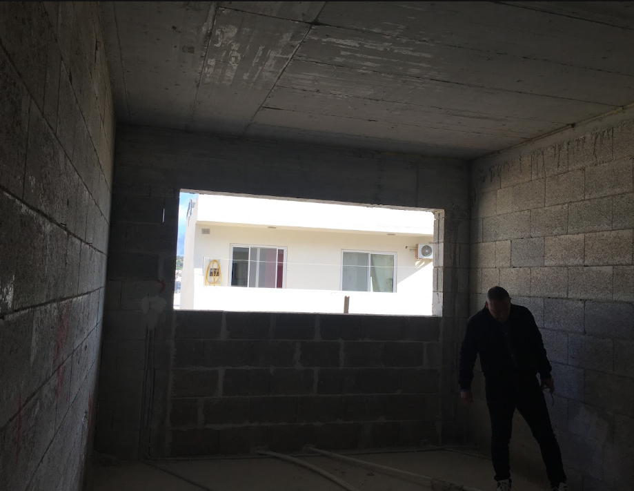 View from inside a concrete building under construction, looking out through a large rectangular opening at a neighboring building with white walls and windows.