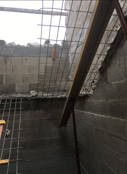 Interior view of a construction site showing unfinished cinder block walls, a partially broken concrete ceiling, metal rebar, and a wooden support beam.