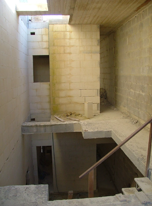 Interior of an unfinished building with gray cinder block walls, exposed concrete floors, and a partial staircase with a metal handrail.