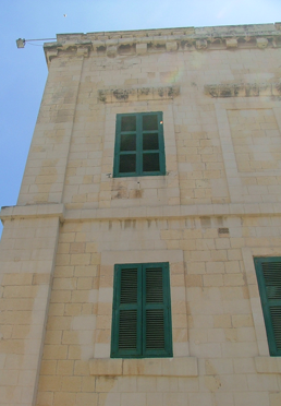 Close-up of a beige stone building with three green window shutters and one green window, against a clear blue sky.